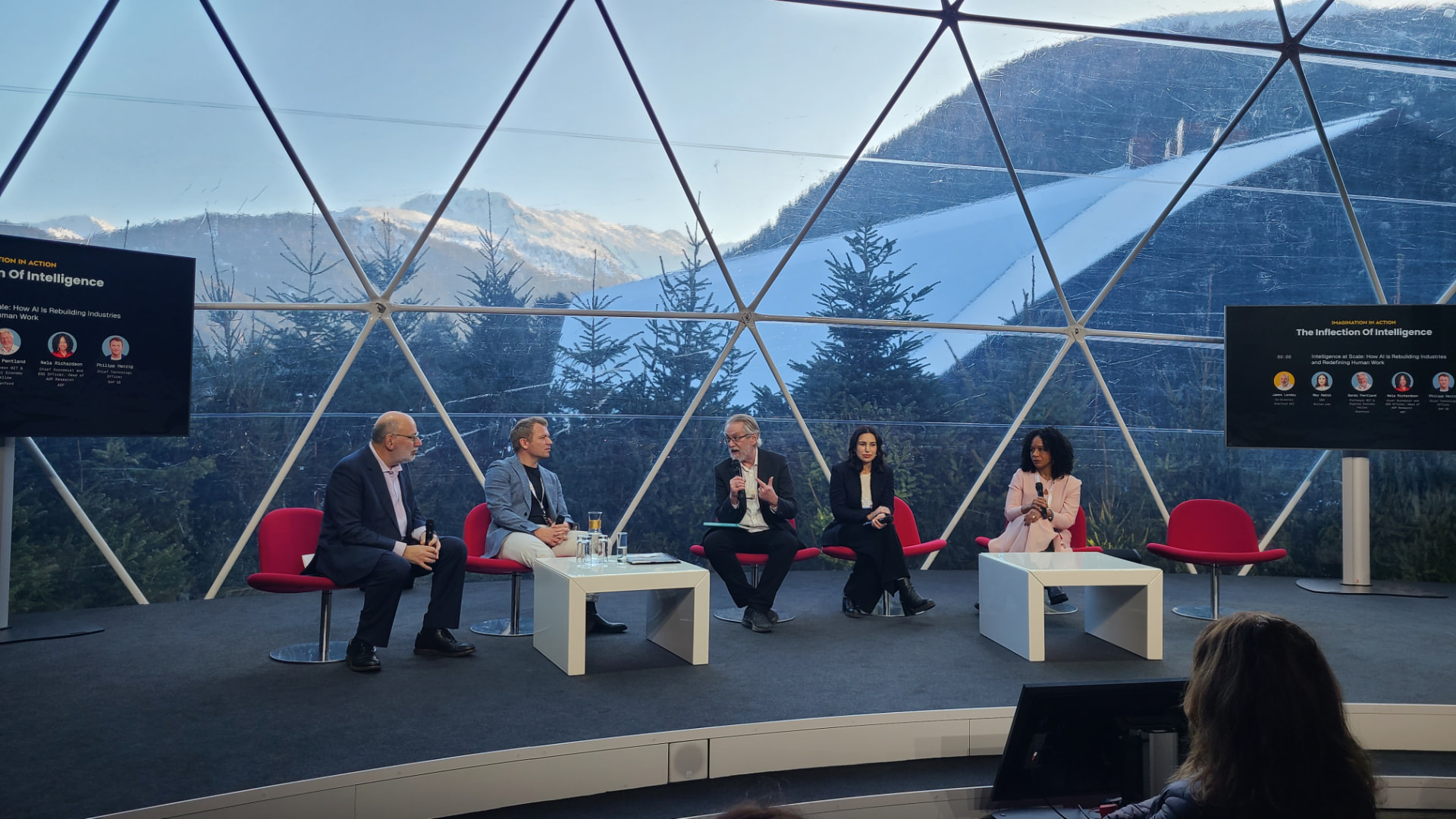 A five-person panel on stage in a glass dome with mountains behind.